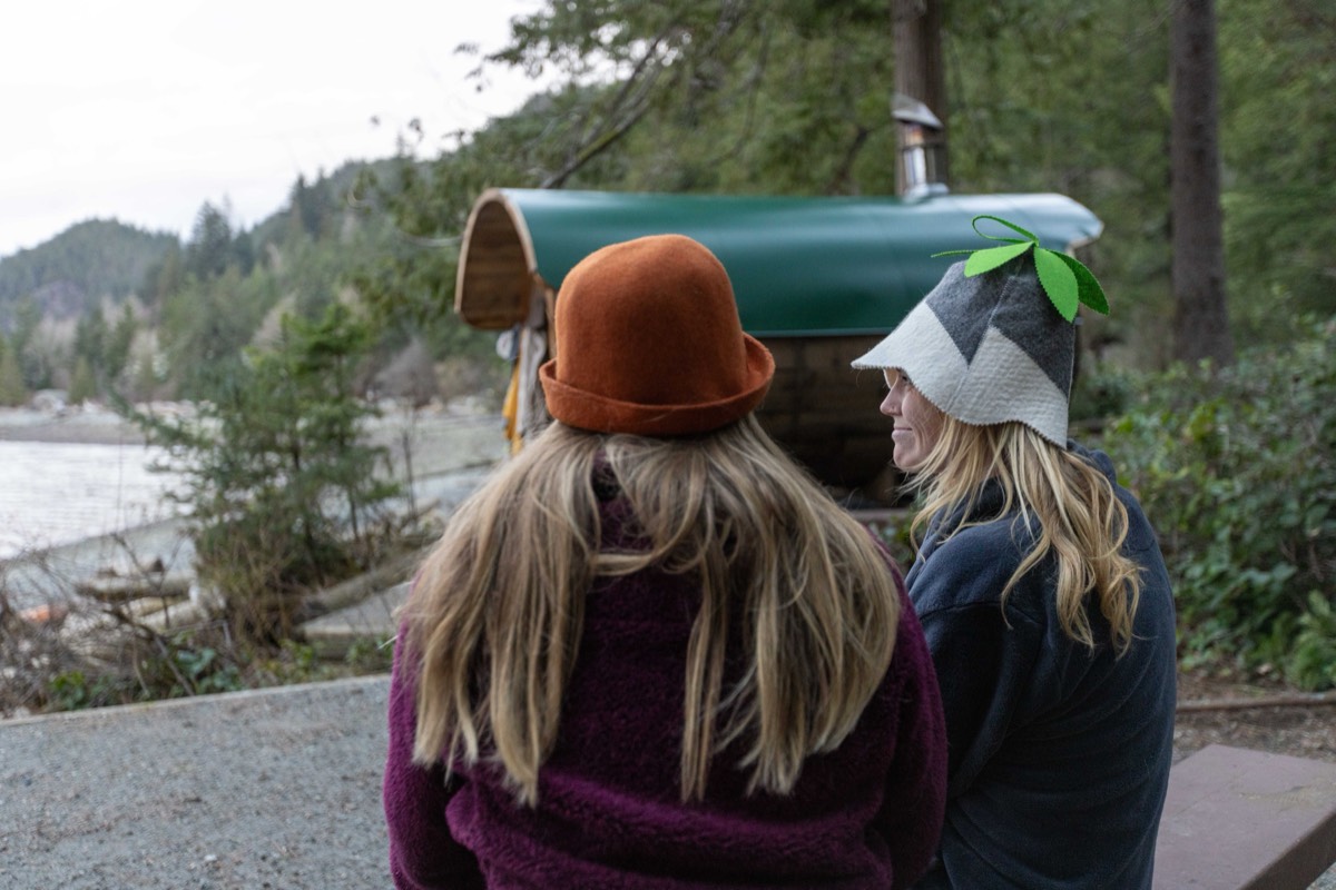 Two friends in felt sauna hats sitting beside the barrel sauna by the water